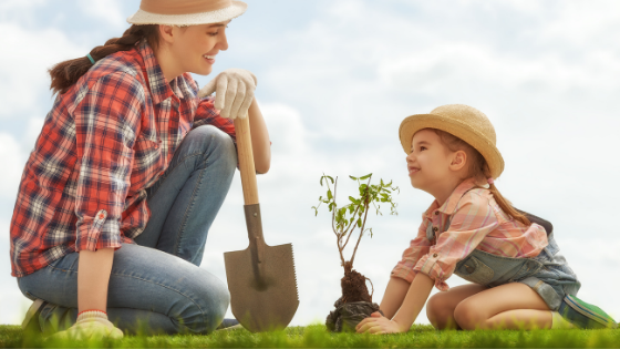 Uma mulher agachada segurando uma pá e uma criança abaixada segurando uma planta.