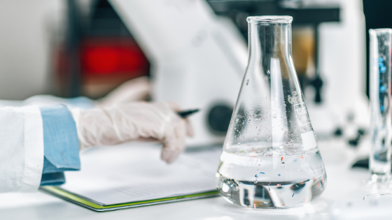 Mão de uma pessoa com luva segurando uma caneta e um caderno e um erlenmeyer na frente.